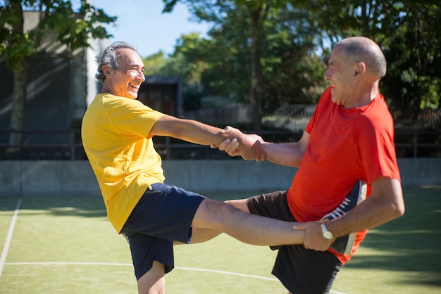 Foto grátis homens sênior úteis se preparando para o jogo em dia ensolarado. homens com cabelos grisalhos em roupas esportivas aquecendo no campo esportivo, ajudando uns aos outros no alongamento. futebol, esporte, conceito de atividades de lazer