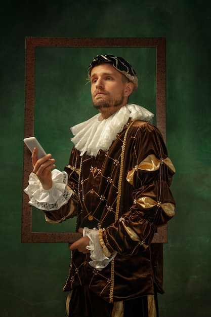 Hora da selfie. Retrato de jovem medieval em roupas vintage, com moldura de madeira em fundo escuro. Modelo masculino como duque, príncipe, pessoa real. Conceito de comparação de eras, moderno, moda.