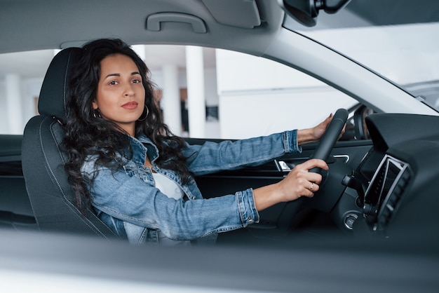 É hora de dar uma volta. Linda garota com cabelo preto experimentando seu novo carro caro no salão automóvel