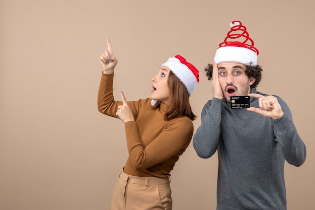 Humor de natal com casal legal animado usando chapéus vermelhos de papai noel chateado cara mostrando o cartão do banco. mulher apontando para cima