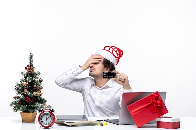 Humor de natal com jovem barbudo exausto empresário com chapéu de papai noel segurando um cartão do banco e apontando para cima no escritório