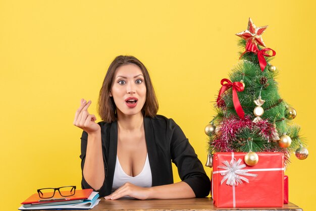 Humor de Natal com jovem mulher bonita concentrada fazendo gestos de dinheiro e sentada no escritório