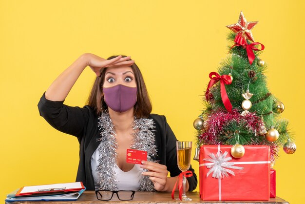 Humor de Natal com mulher bonita concentrada de terno usando máscara médica segurando o cartão do banco no escritório em amarelo