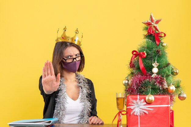 Humor de natal com uma bela dama de terno usando coroa e máscara médica fazendo gesto de parada no escritório em amarelo