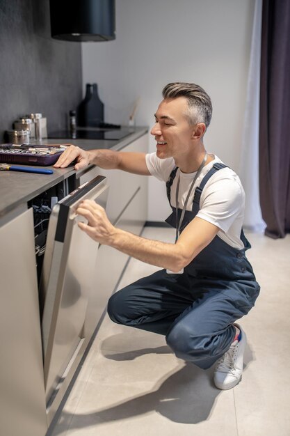 Humor de trabalho Homem sorridente espiando pela porta aberta da máquina de lavar louça embutida na cozinha moderna