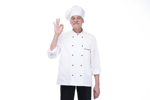 Idoso chef profissional sênior, posando com o solteiro ok, isolado sobre uma parede branca