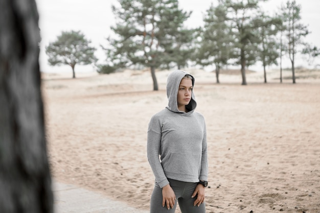 Imagem ao ar livre de jovem esbelta em forma vestida com roupas esportivas elegantes, posando do lado de fora com a praia de areia e pinheiros no fundo, fazendo exercícios, fazendo a rotina de exercícios matinais. foco seletivo