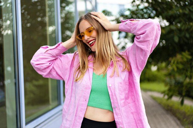 Imagem ao ar livre de uma mulher loira bonita e elegante, posando perto de uma janela urbana espelhada, roupa brilhante de blogueira e maquiagem.