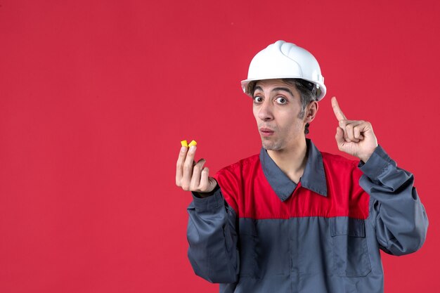 Imagem aproximada de um jovem construtor chocado, de uniforme, com capacete e tampões de ouvido apontando para cima na parede vermelha isolada