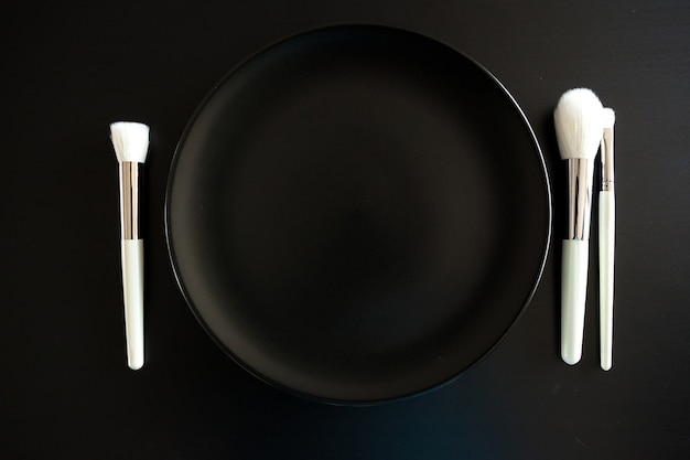 Imagem conceitual de pincéis de maquiagem ao lado do prato de jantar em fundo preto