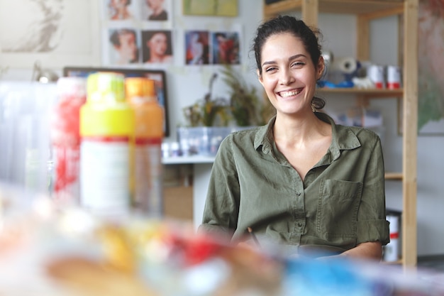 Imagem da charmosa artista feminina carismática, vestindo camisa cáqui, sorrindo amplamente sentindo-se feliz com seu trabalho e processo de criação, sentada na oficina, rodeada de acessórios de pintura