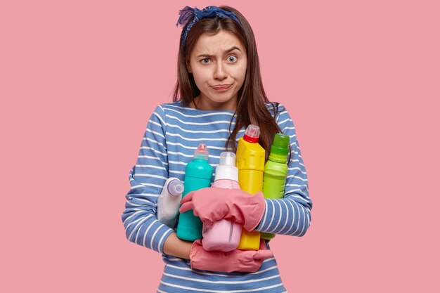 Foto grátis imagem da cintura para cima de uma jovem frustrada e descontente segurando frascos de diferentes produtos de limpeza, franzindo os lábios em descontentamento