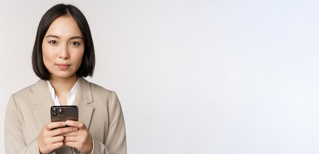 Imagem da empresária asiática de terno segurando o celular usando o aplicativo de smartphone sorrindo para a câmera de fundo branco