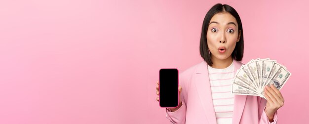 Imagem da mulher corporativa bem sucedida coreana mostrando dólares de dinheiro e interface de tela de aplicativo de smartphone do conceito de aplicativo de telefone móvel de investimento e finanças fundo rosa