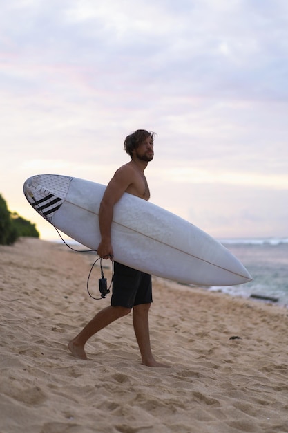 Imagem da paisagem do surfista masculino ocupado andando na praia ao nascer do sol enquanto carregava sua prancha debaixo do braço com as ondas do mar quebrando no fundo. Jovem surfista masculino bonito no oceano