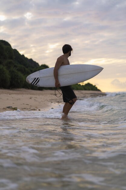 Imagem da paisagem do surfista masculino ocupado andando na praia ao nascer do sol enquanto carregava sua prancha debaixo do braço com as ondas do mar quebrando no fundo. Jovem surfista masculino bonito no oceano