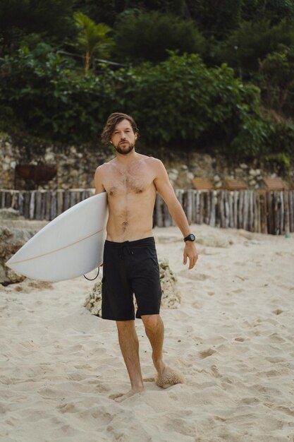 Imagem da paisagem do surfista masculino ocupado andando na praia ao nascer do sol enquanto carregava sua prancha debaixo do braço com as ondas do mar quebrando no fundo. Jovem surfista masculino bonito no oceano