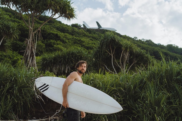 Imagem da paisagem do surfista masculino ocupado andando na praia ao nascer do sol enquanto carregava sua prancha debaixo do braço com as ondas do mar quebrando no fundo. jovem surfista masculino bonito no oceano