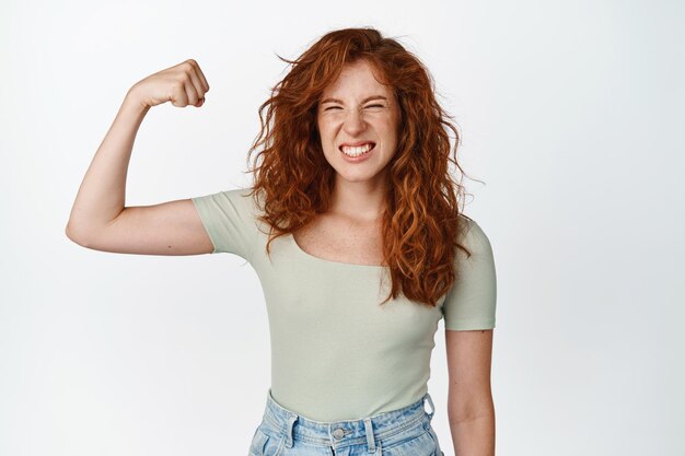 Imagem de adolescente ruiva atrevida flexionando bíceps mostrando músculo no braço e parecendo feliz gesto de poder feminino se sentindo forte em pé sobre fundo branco