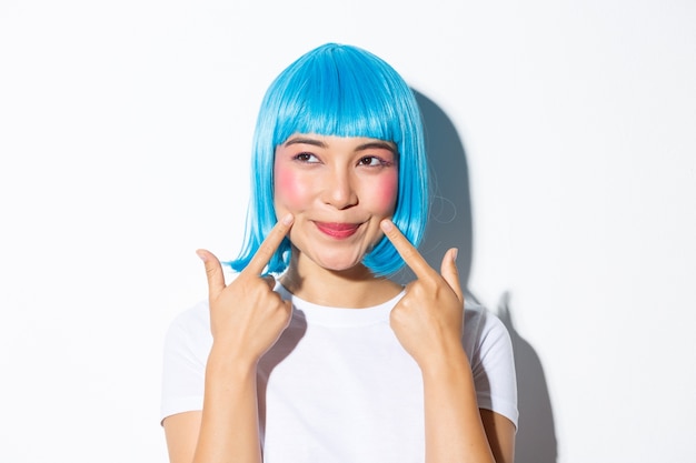 Imagem de adorável garota asiática com peruca azul cutucando suas bochechas e olhando o canto superior esquerdo, em pé.