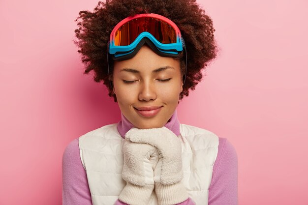 Imagem de close-up de uma jovem encaracolada relaxou com uma expressão de rosto feliz, mantém as mãos com luvas brancas pressionadas juntas, usa equipamento de snowboard, fecha os olhos e sorri suavemente, aproveita o inverno