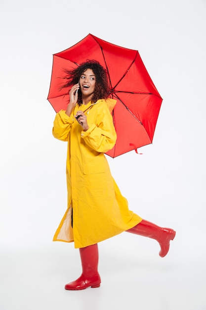 Imagem de corpo inteiro de surpresa mulher africana feliz em capa de chuva se escondendo sob o guarda-chuva e andando no estúdio enquanto fala pelo smartphone e olhando para trás sobre fundo branco