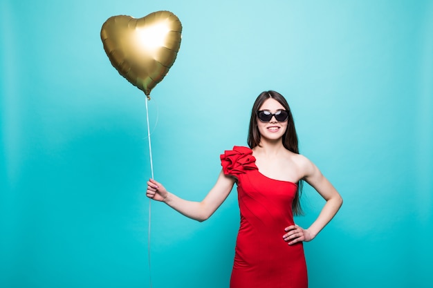 Imagem de corpo inteiro de uma mulher linda em um traje vermelho chique posando com um balão em forma de coração, isolado sobre uma parede verde