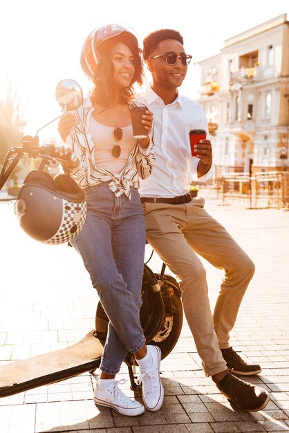 Imagem de corpo inteiro do feliz casal Africano jovem bebendo café em pé perto da moto moderna na rua e olhando para longe