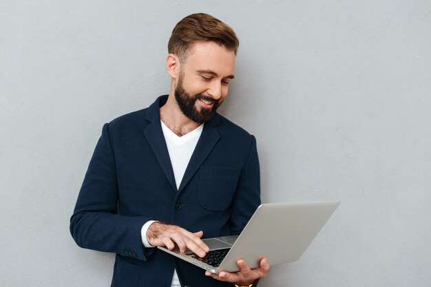 Imagem de corpo inteiro do homem barbudo sorridente em roupas de negócios usando o computador portátil sobre cinza