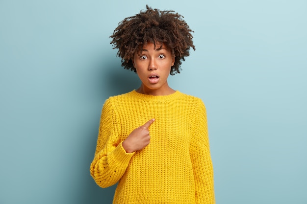 Imagem de espantada indignada surpresa jovem com cabelo afro encaracolado, sem fala, aponta para si mesma, usa suéter amarelo