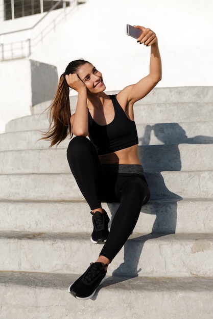 Imagem de esportes linda mulher sentada nos degraus ao ar livre e olhando de lado faz selfie por telefone.