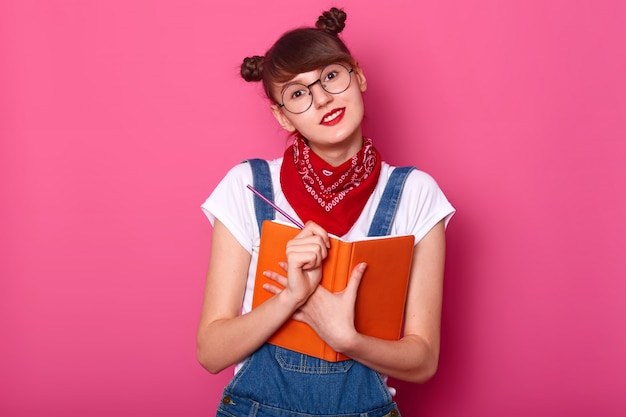 Foto grátis imagem de estúdio de adolescente feminino bonito com dois cachos engraçados, segurando o diário e o lápis nas mãos, olhando diretamente