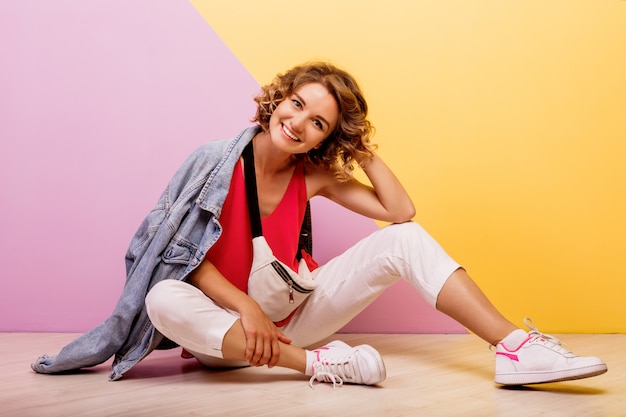 Imagem de estúdio de mulher adorável morena sorridente, vestindo roupa desportiva elegante e jaqueta jeans, sentada no chão.