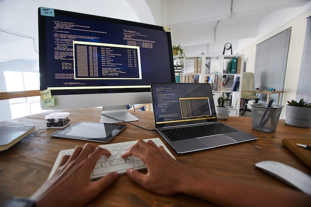 Imagem de fundo de mãos masculinas digitando no teclado com código de programação preto e laranja na tela do computador e laptop no interior contemporâneo do escritório, copie o espaço