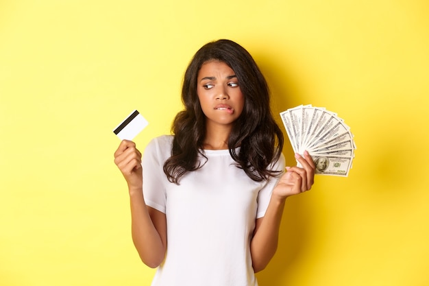 Imagem de garota afro-americana indecisa fazendo escolha entre dinheiro e cartão de crédito