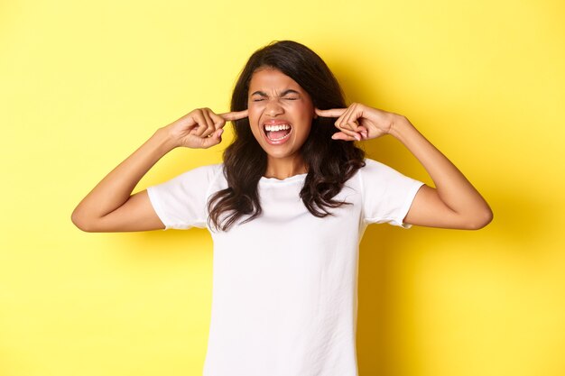 Imagem de garota afro-americana irritada, não suporto barulho incomodando alto, ouvidos fechados e gritando irritada, em pé sobre fundo amarelo.