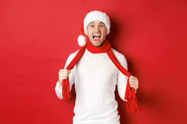 Imagem de homem adulto com raiva odeio natal usando lenço e chapéu de Papai Noel gritando angustiado em pé louco ...