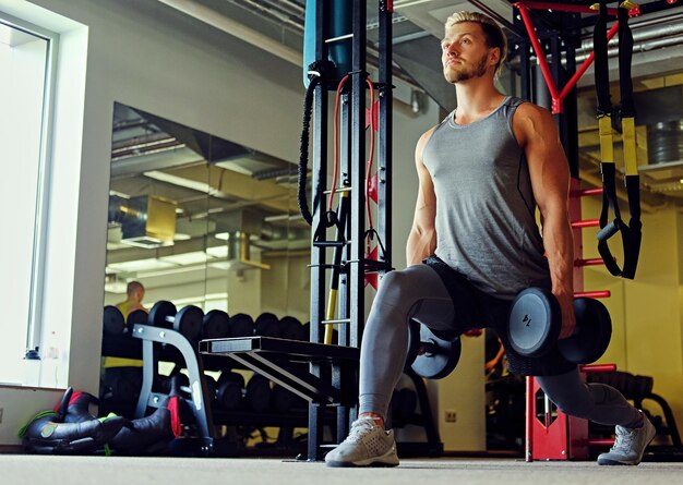 Imagem de homem atlético de corpo inteiro fazendo agachamentos com halteres em um clube de ginástica.