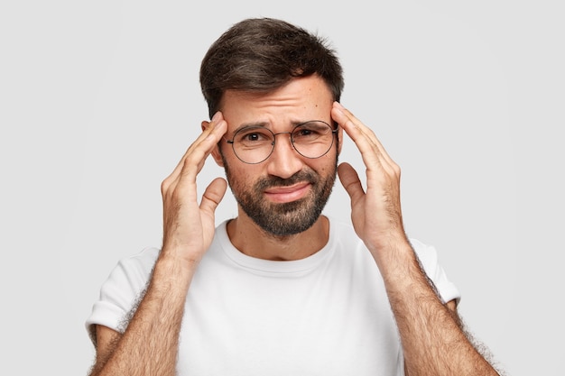 Imagem de homem barbudo descontente sofre de forte dor de cabeça depois de trabalhar a noite toda, tem expressão de fadiga, mantém as mãos nas têmporas, franze a testa, posa contra uma parede branca. mau pressentimento