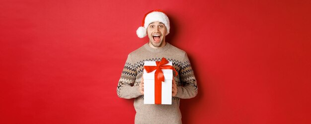 Imagem de homem bonito animado recebendo presente de natal, usando chapéu de papai noel e suéter de inverno, gritando de alegria, segurando o presente e de pé sobre fundo vermelho