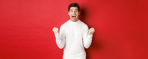 Imagem de homem bonito feliz de suéter branco, ganhando algo, fazendo punho bomba e sorrindo espantado, comemorando a vitória, de pé sobre fundo vermelho.