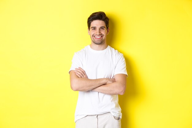 Imagem de homem caucasiano confiante sorrindo satisfeito, de mãos cruzadas no peito e parecendo satisfeito, em pé sobre um fundo amarelo.