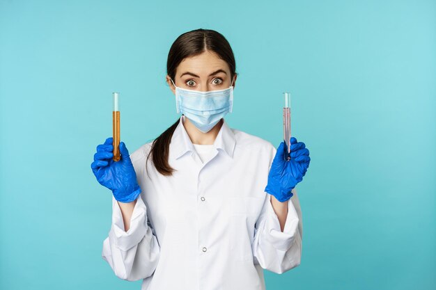 Imagem de jovem médica, trabalhadora de laboratório fazendo pesquisa, segurando tubos de ensaio, usando máscara facial médica e luvas de borracha, fundo azul