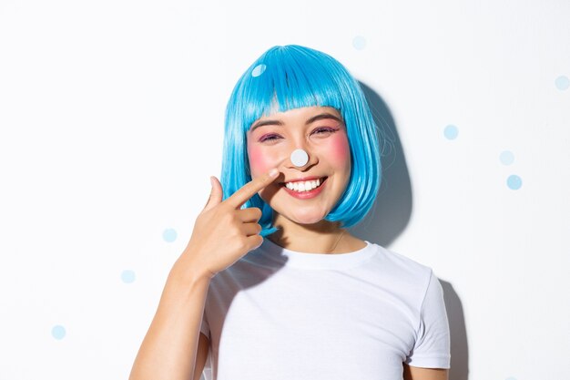 Imagem de linda e boba garota asiática com confete no nariz, sorrindo e parecendo feliz, usando peruca azul para festa de halloween.