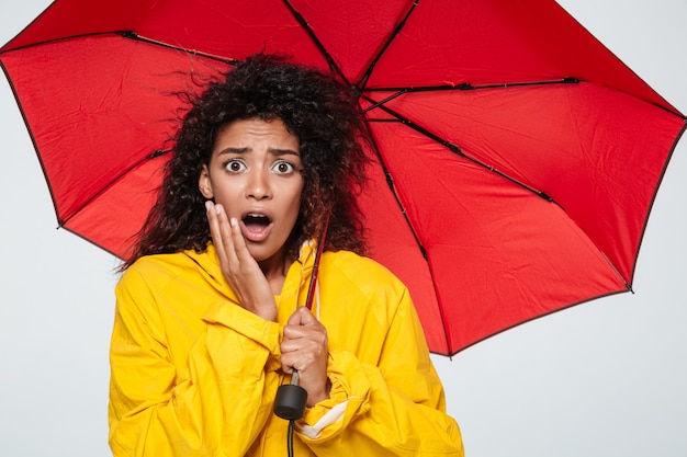 Imagem de mulher africana chocada na capa de chuva se escondendo sob o guarda-chuva