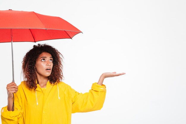 Imagem de mulher africana confusa na capa de chuva se escondendo sob o guarda-chuva e esperando raing sobre branco