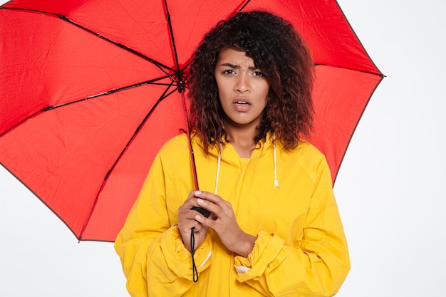 Imagem de mulher africana confusa na capa de chuva se escondendo sob o guarda-chuva
