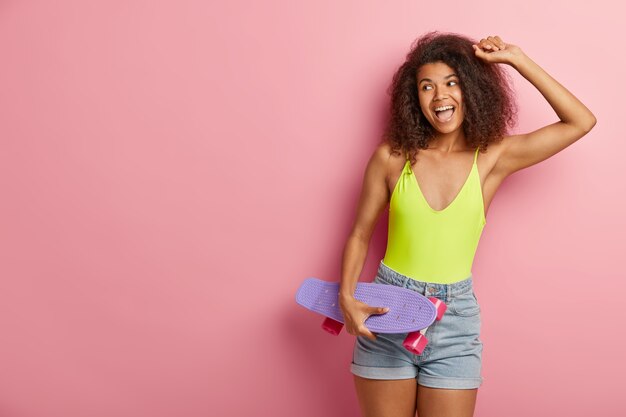 Imagem de mulher afro otimista e despreocupada com pele escura, dançando ativamente com o skate, se divertindo em dia ensolarado