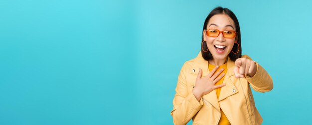 Imagem de mulher coreana feliz em óculos de sol, apontando o dedo para a câmera com expressão de rosto surpreso e alegre surpreso em pé sobre fundo azul