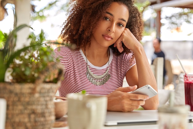 Foto grátis imagem de mulher negra séria de etnia lê mensagem recebida no celular, verifica e-mail no celular, senta-se no interior do café com uma caneca de café, conectada à internet sem fio, faz compras